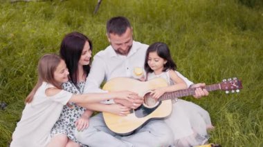 Handsome man playing guitar for his charming wife and two cute daughters. Happy caucasian family sitting on soft blanket at garden and singing songs. Parents and kids spending leisure time together.