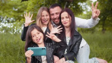 Positive family sitting together on soft plaid utdoors and taking selfie on modern smartphone. Young parents with two kids saving memories. Concept of people, leisure and technology