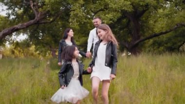 Ebeveynleri el ele tutuşup arka planda yürürken iki neşeli kız kardeş birlikte yeşil bahçede oynuyorlar. Dört kişilik mutlu bir aile temiz havanın tadını çıkarıyor. İki çocuklu aile.