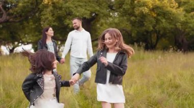 İki çocuklu pozitif aile yaz günlerini yeşil bahçede geçiriyor. Mutlu kız kardeşler bulanık ön planda eğleniyorlar. Genç ebeveynler yürür ve arkada konuşurlar. Oynayan kız kardeşler.