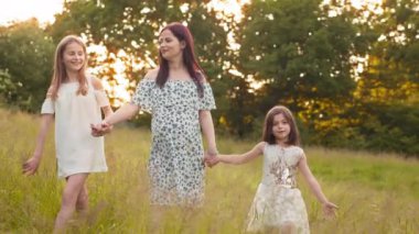 Attractive pregnant mother holding hands with her two little daughters walking at green garden. Young woman spending summer days with kids on fresh air. Pregnant woman with two daughters posing.