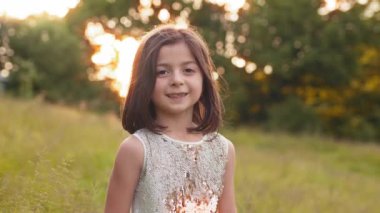 Portrait of pretty little girl with short brown hair posing at green garden with beautiful sunrise behind. Concept of happiness and childhood. Portrait of pretty girl with brown hair posing at garden.