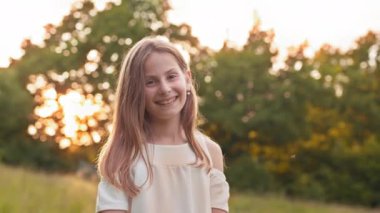 Portrait of cute girl in white summer dress smiling and looking at camera while standing at green garden. Charming little lady with blond hair enjoying warm days outdoors. Girl in summer dress.