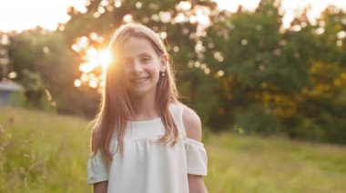 Pretty young girl wearing stylish white dress posing at green garden. Beautiful summer sunrise on background. Concept of childhood and carefree. Cute girl smiling and looking at camera at garden.