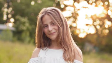 Attractive young blonde teen girl walking in summer sunset park, looks at camera with alluring look. Portrait of a young woman outdoors. Happy little girl, Children in park.