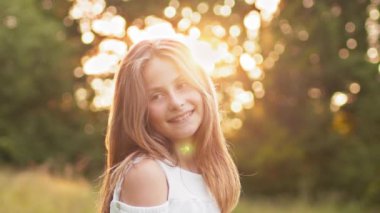 Pretty blond teenage girl with wavy hair smiling on camera looking cheerful. Positive emotions. Attractive young blonde teen girl walking in summer sunset park, looks at camera with alluring look