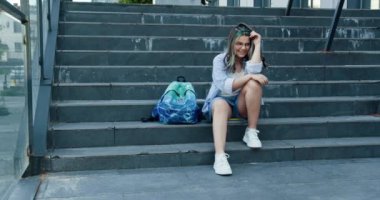 Dışarıda taş merdivenlerde oturan genç kız yanında sırt çantasıyla gülümsüyor. Beyaz, kadın, okul temalı, neşeli ifadeli günlük kıyafetler. Rahat ve tasasız bir atmosfer..