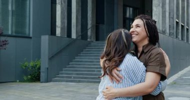 Farklı geçmişlere sahip iki kadın modern binanın dışında kucaklaşıyorlar, neşe ve dostluğu ifade ediyorlar. Orta yaşlı beyaz kadın ve genç siyah kadın mutlu anlarını paylaşıyorlar..