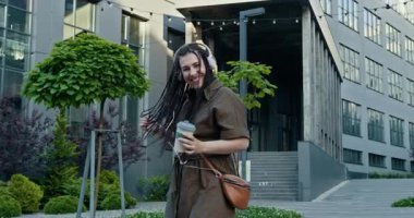 Kulaklıklı neşeli bir kadın yeşilliklerle dolu canlı bir şehir manzarasında mutlu bir şekilde dans ediyor. Neşeli bir kadın açık havada parlak kulaklıklarla dans ediyor, müziğine ve mutluluğuna dalmış.