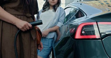 Şehirde iki genç kadın elektrikli araba şarj ediyor. Bir tanesi şarj kablosu tutuyor. Çeşitli modeller sürdürülebilirliği ve modern yaşam tarzını vurgular. Anne ve kızı şehirde elektrikli araba şarj ediyorlar.