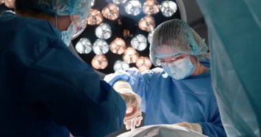 Mavi ameliyat önlüklü iki cerrah parlak ışıklar altında ameliyat yapıyor. Maske ve şapka takan yetişkin profesyoneller hassasiyet ve özen üzerine odaklanırlar. Modern tıp ortamında takım çalışması.