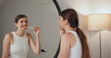 Genç beyaz kadın banyoda dişlerini fırçalıyor. Aynadaki yansıma, beyaz diş fırçası ve günlük kıyafetlerle dişçi temizliği rutinini gösteriyor. Yeni ve temiz bir sabah atmosferi yaratıyor..