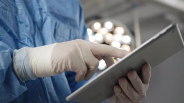 Steril eldiven ve önlük giyen bir tıp uzmanı ameliyat odasında muhtemelen tıbbi veriler için bir tablet kullanır. Bir doktor ya da cerrah, maske ve ameliyat önlüğü takıyor.