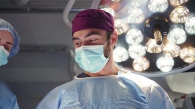 Ameliyat önlüğü ve yüz maskesi takan iki tıp uzmanı, muhtemelen bir ameliyathanede, biri kepli olmak üzere cerrahi ışıklarla aydınlatılıyor. Tıbbi ekip ameliyattan sonra beşlik çakıyor.