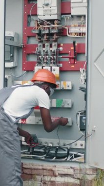 An electrician wearing a hard hat and safety glasses inspects an electrical panel with a multimeter, ensuring safety and functionality.