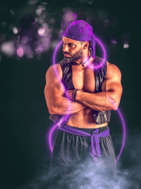 Güçlü gövdesi olan, parlak mor ışıkları olan ve kolları karanlıkta sihirli bir cin gibi katlanmış yakışıklı yetişkin bir erkek.