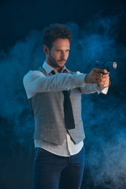 Stüdyoda yakışıklı bir adam silah doğrultuyor, şık kıyafetler giyiyor, koyu bir arka planı var.
