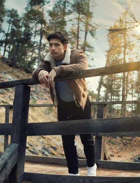秋や冬には山の中で魅力的な若い男が木製の橋にもたれ スエードのジャケットを着て カメラを見て — ストック写真