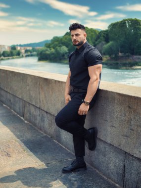 Nehir kıyısında yürüyen çekici atletik adam siyah gömlek ve pantolon giyiyor.