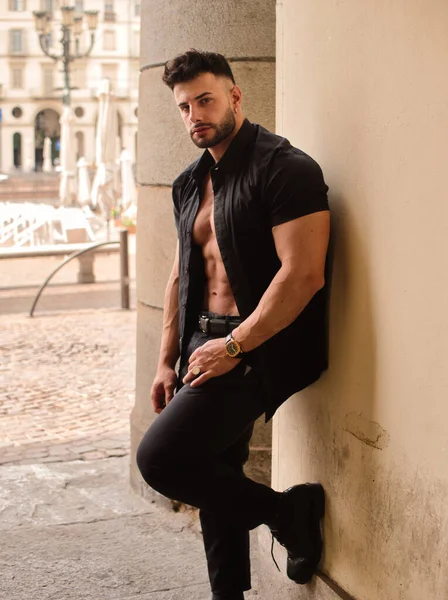 Attractive athletic man with shirt open on naked muscular torso, leaning against pillar, in European city center, wearing black shirt and pants