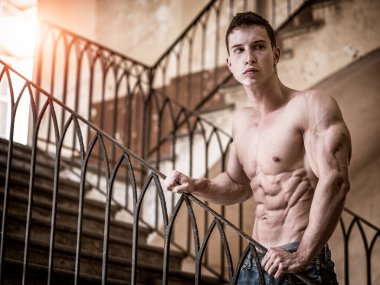 Üstsüz, kaslı, kotlu genç bir adam. Kapalı alanda duruyor. Gövde kaslarını gösteriyor.