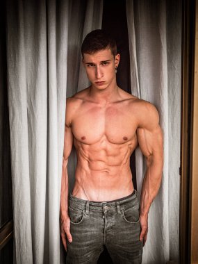Üstsüz, kotlu yakışıklı genç adam, iç mekan da duruyor, vücut kaslarını gösteriyor, perdelerin yanında, kameradan dışarı bakıyor.