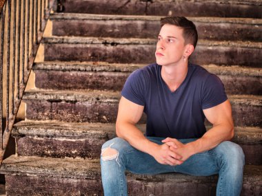 Yakışıklı yakışıklı genç adam merdivenlerde oturuyor, kaslı kollarını gösteriyor, tişört ve kot pantolon giyiyor.