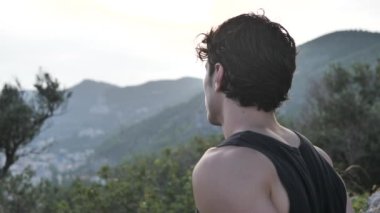 Tanktop 'u olan ağaçların arasındaki yakışıklı genç adam gün batımında güneş ışığında dinleniyor ve kameraya bakıyor.