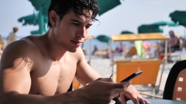 运动的年轻人在海边使用手机键入信息 而看着海和海滩在日落 — 图库视频影像