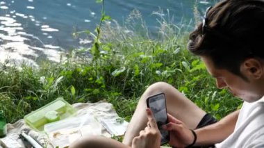 Genç adam, doğada piknik yaparken akıllı telefonuyla yemek fotoğrafı çekiyor.