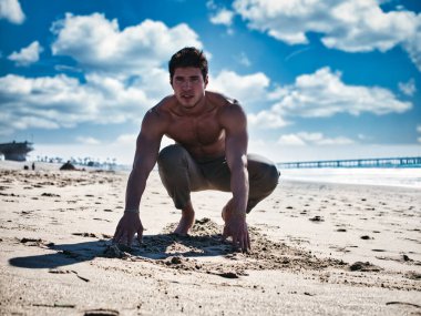 Handsome young man on beach in a sunny day, sitting on sand, looking at camera