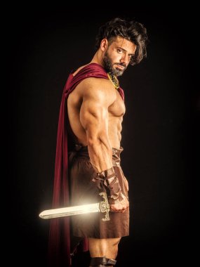Handsome muscular man posing in roman or spartan gladiator costume with shield and sword, isolated on black background in studio