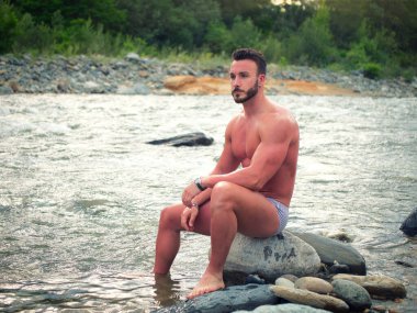 Attractive muscular shirtless young man in nature next to a river, wearing swimming suit, sitting on rock