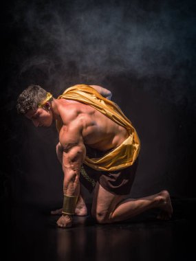 Serious young man in Greek god or Roman aristocrat costume with muscular body, with fist against the ground, looking down on black background