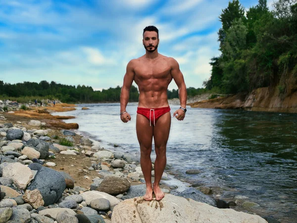 Attractive muscular shirtless young man in nature next to a river, wearing swimming suit, standing