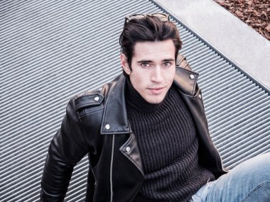 Metal şebekede oturan yakışıklı genç adam şehir ortamında kameraya bakıyor.