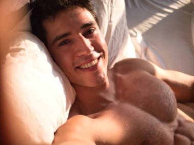 Attractive young man smiling at cell phone to take selfie photo, while laying on bed, shirtless