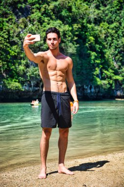 Full body shot of a handsome young man using cell phone to take selfie photo, standing on a beach, shirtless wearing boxer shorts, showing muscular fit body