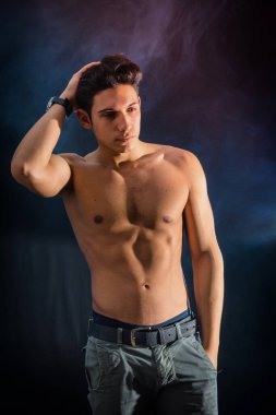 Three quarters shot of handsome shirtless athletic young man in pants, looking away in studio shot, on dark smoke background