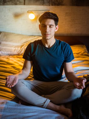 Genç ve çekici bir adam yatağında meditasyon yapıyor, lotus pozisyonunda gözleri kapalı oturuyor ve sağlık ve zindelik kavramında sükunet ifadesi sergiliyor.