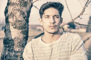 Kırsalda yakışıklı, zinde bir genç adam duruyor ve ağaca yaslanıyor, başka tarafa bakıyor.