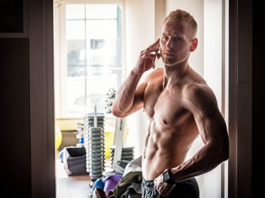 Spor salonunda cep telefonu kullanan kaslı sarışın genç adam birini arıyor.