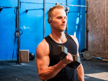 Yakışıklı sarışın, bisepsi kaldıran, spor salonunda duran, bir tarafa bakan genç adam.