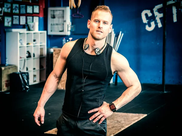 Handsome muscular young man standing in a gym, looking at camera, with headphones around his neck