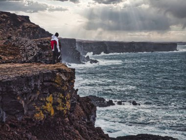 İzlanda 'da fırtınalı bir günde dalgalı kayalıkların yakınındaki kayalara çarpan deniz suyuna bakan bir adam.
