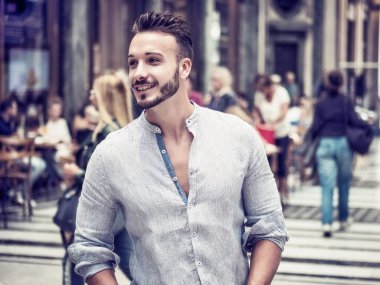 Avrupa şehrinde yakışıklı bir genç adam ayakta, tişörtünü giymiş ve gülümsüyor.