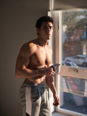 Seksi yakışıklı genç adam yatak odasında üstsüz duruyor pencere perdelerinin yanında elinde kahve fincanı ya da fincanı var ve kameraya bakıyor.