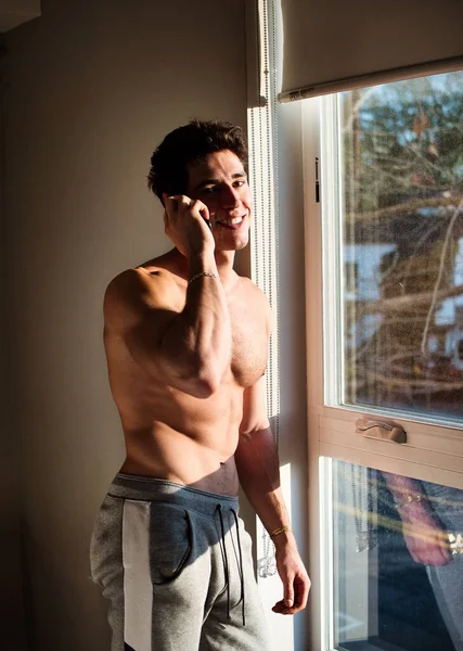 ハンサムな若い男は 窓の隣の彼の寝室で 携帯電話でシャツなしで話している — ストック写真
