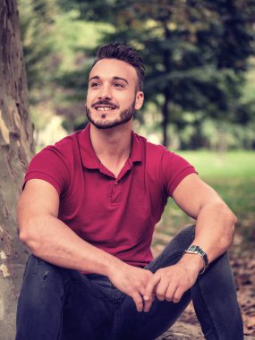 Güneşli bir yaz gününde parkta dinlenen ya da ağaca yaslanan çekici genç bir adam.