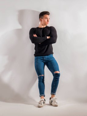 Stüdyoda yakışıklı bir genç adam, beyaz duvara karşı duruyor, kollarını kavuşturmuş, siyah kazak ve kot pantolon giyiyor.